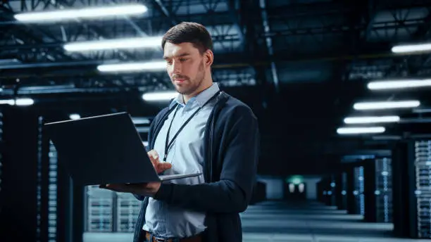 Photo of Successful Data Center IT Specialist Using Laptop Computer. Server Farm Cloud Computing Facility with System Administrator Working. Data Protection Engineering Network for Cyber Security.