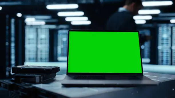 Photo of Laptop Computer with Green Screen Chroma Key Mock Up Display Stands on a Table. In the Background Data Center With Rows of Server Racks and IT Engineer Working. Advanced Equipment.