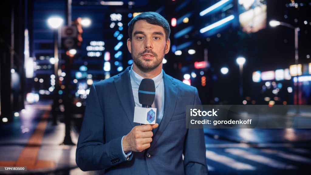 Anchorman Reporting Live News in a City at Night. News Coverage by Professional Handsome Reporter from a Business District. Journalist Presenting News for TV Channel. Newscaster Talking. Journalist Stock Photo