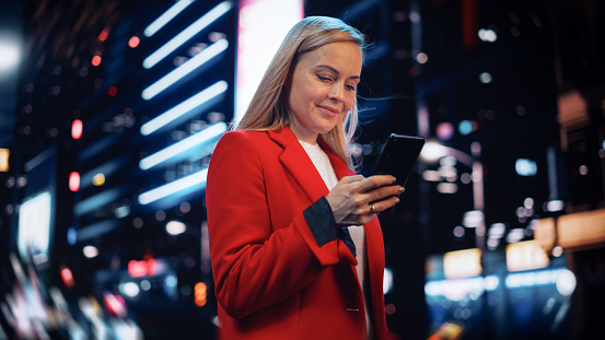 Beautiful Woman Standing, Using Smartphone on City Street with Neon Bokeh Lights Shining at Night. Confident Smiling Beautiful Female Using Mobile Phone. Low Angle Medium Shot Cinematic Portrait.