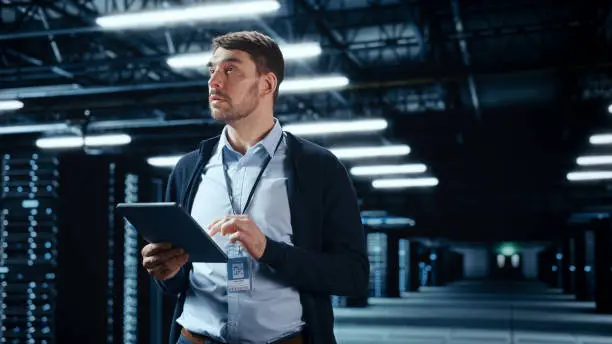 Photo of Successful Data Center IT Specialist Using Tablet Computer. Server Farm Cloud Computing Facility with System Administrator Working. Data Protection Engineering Network for Cyber Security.