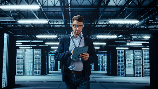 Male IT Specialist Walks Between Row of Operational Server Racks in Data Center. Engineer Uses Tablet Computer for Maintenance. Concept for Cloud Computing, Artificial Intelligence, Cybersecurity.