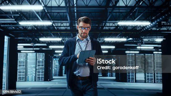 istock Male IT Specialist Walks Between Row of Operational Server Racks in Data Center. Engineer Uses Tablet Computer for Maintenance. Concept for Cloud Computing, Artificial Intelligence, Cybersecurity. 1394184402