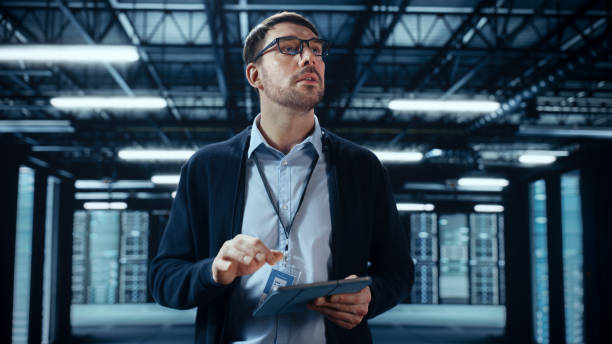 istock Male IT Specialist Walks Between Row of Operational Server Racks in Data Center. Engineer Uses Tablet Computer for Maintenance and Diagnostics in Cyber Security and Data Protection Facility. 1394184435