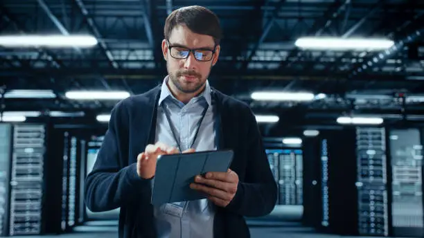 Photo of Male IT Specialist Walks Between Row of Operational Server Racks in Data Center. Engineer Uses Tablet Computer for Maintenance. Concept for Cloud Computing, Artificial Intelligence, Cybersecurity.