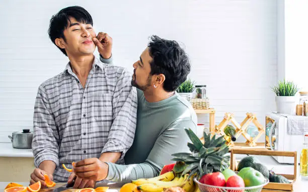 Gay LGBT sweet Asian couple wearing pajamas, smiling, teasing and pinching cheek with happiness and love, eating healthy fruits for breakfast in kitchen at home in the morning. Lifestyle Concept