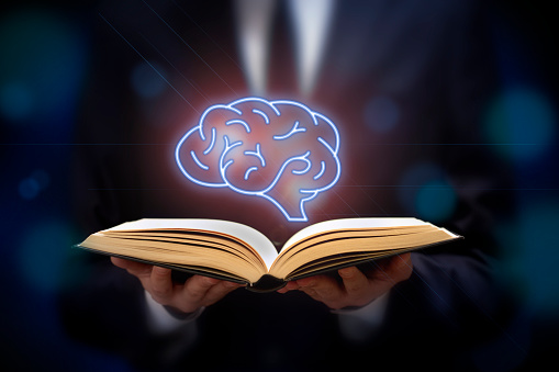 Front view of two human hands holding an opened book with a floating brain on top. Predominant color is dark blue.