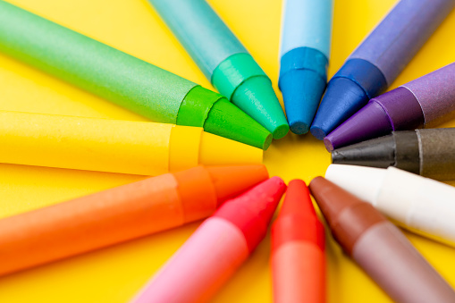 Top view of various crayons arranged side by side on a multicolored gradient circle on yellow background