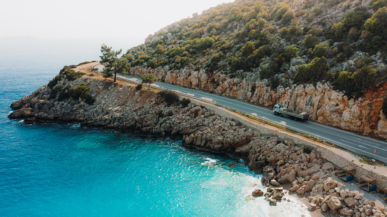 Drone panoramic photo of a car driving the highway road surrounded by the crystal blue sea, mountains and the island view in Antalya region, South Turkey
