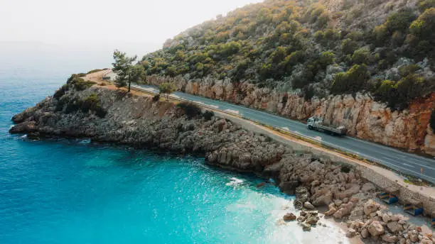Drone panoramic photo of a car driving the highway road surrounded by the crystal blue sea, mountains and the island view in Antalya region, South Turkey