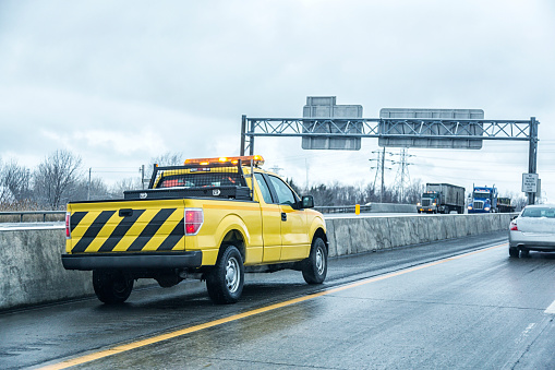 A bright yellow, black striped tailgate, roadside emergency or maintenance vehicle pickup truck is parked - waiting on the far left inside shoulder of a multiple lane expressway - on a slick, slippery winter road day during a light snow storm in western New York State in early February.