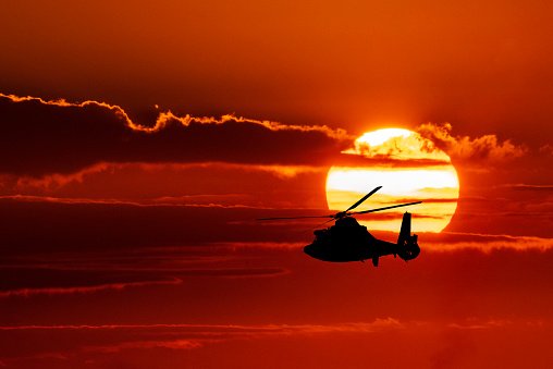 A U.S. Coast Guard helicopter flies in silhouette against the setting sun in a composite image.