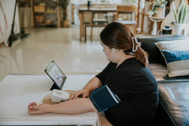 Telemedicine, Asian plus size woman checking her blood and heart rate pressure by herself at home. Asian woman checking her blood pressure while having virtual appointment with her doctor. pudgy woman pictures stock pictures, royalty-free photos & images