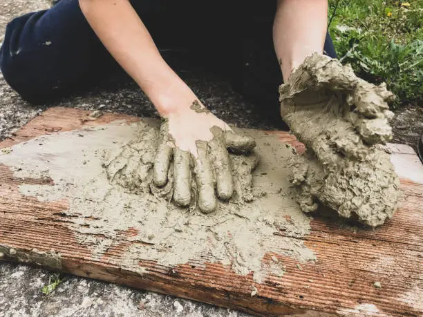 Photo of A kid playing with wet sand. A kid kneads and models wet clay on a wooden plank on the floor. Wet sand games.