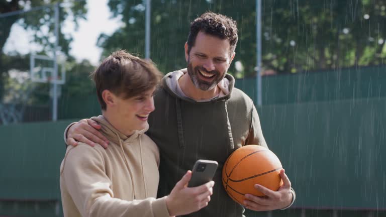 Father and son using a smartphone after playing a game of basketball. Young man and teenage boy having fun while staying fit, active