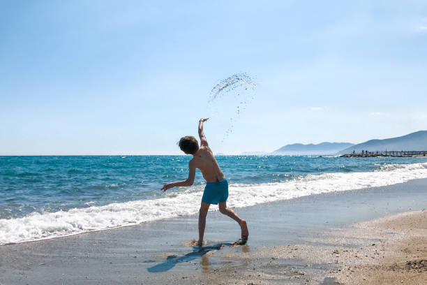 A little boy plays on the beach by the clear blue sea throwing sand in the air in the summer on a beautiful sunny day with blue sky. Liguria, Italy. A little boy plays on the beach by the clear blue sea throwing sand in the air in the summer on a beautiful sunny day with blue sky. Liguria, Italy. varigotti stock pictures, royalty-free photos & images