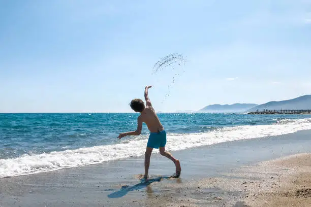 A little boy plays on the beach by the clear blue sea throwing sand in the air in the summer on a beautiful sunny day with blue sky. Liguria, Italy.