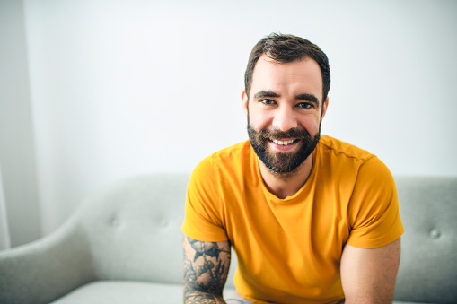 A portrait of casual young man with beard and tattoo and Burnt Orange shirt sit on the sofa at home