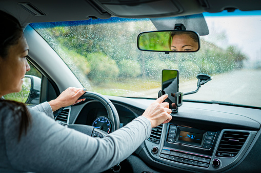 Close up view of a woman touching mobile placed on phone car mount while driving through wet road in a rainy day. High resolution 42Mp outdoors digital capture taken with SONY A7rII and Zeiss Batis 40mm F2.0 CF lens