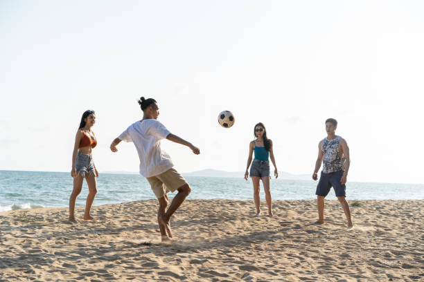 groupe de jeunes hommes et femmes asiatiques jouent au football sur la plage ensemble. les voyageurs amis attrayants se sentent heureux et se détendent, ayant un match de football tout en voyageant pour des vacances de vacances dans l’île de la mer tro - foot walk photos et images de collection