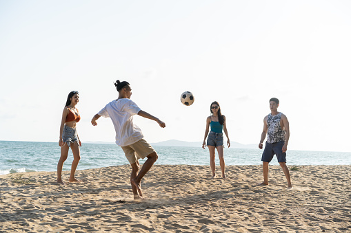 Group of Asian young man and woman play soccer on the beach together. Attractive friend traveler feel happy and relax, having a football game while travel for holiday vacation in tropical sea island.