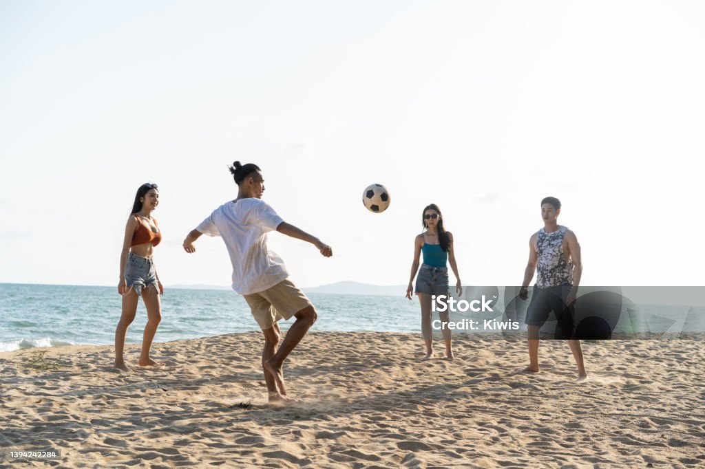 Group of Asian young man and woman play soccer on the beach together. Attractive friend traveler feel happy and relax, having a football game while travel for holiday vacation in tropical sea island. Beach Stock Photo