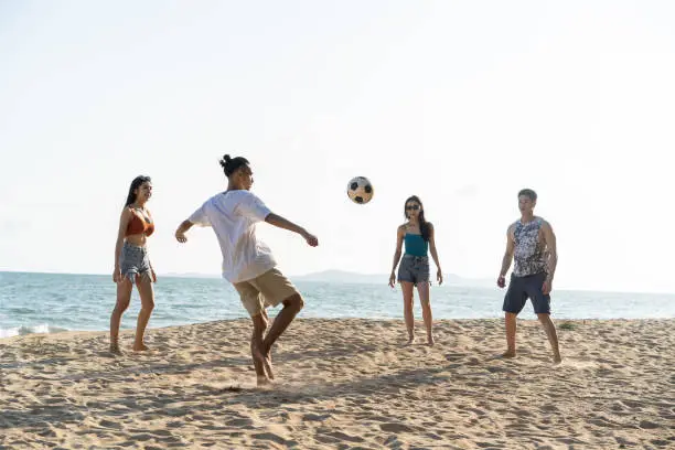 Photo of Group of Asian young man and woman play soccer on the beach together. Attractive friend traveler feel happy and relax, having a football game while travel for holiday vacation in tropical sea island.