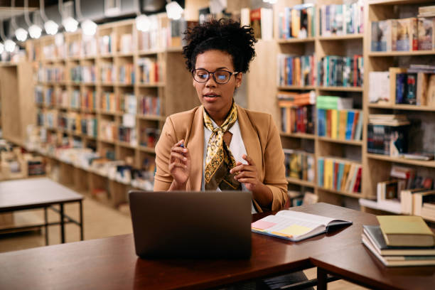 istock Black female teacher talking while holding online class over laptop from a library. 1394244667