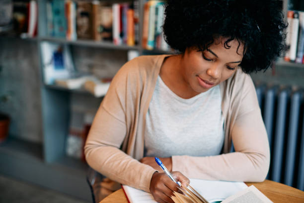 istock Black mid adult woman writing notes while doing a research in library. 1394244781