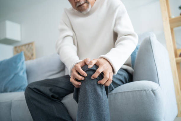 Asian Senior elderly disabled man sit on sofa suffer from injured leg. Mature older male patient sitting on couch in living room has knee problem and feel painful. Insurance and Healthcare concept. stock photo