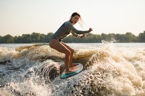 screaming wet blonde woman wakesurfer in grey swimsuit riding on splashing wave on a warm summer day