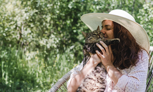 Young adult curly woman in hat embracing with the domestic cat in the garden.