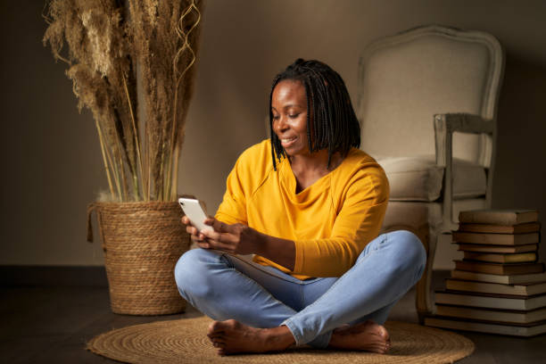 Portrait of a black woman with braided hair sitting on the ground with a mobile phone in her hands stock photo