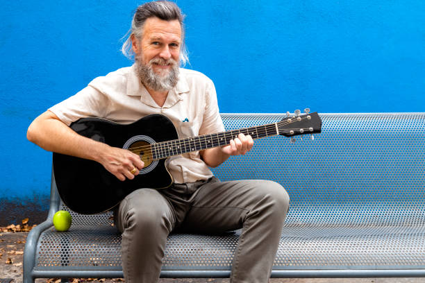 istock Smiling mature caucasian man with beard sitting on a bench playing the guitar looking at camera. Copy space. 1394257842