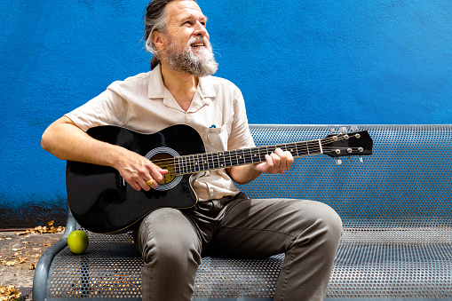 Mature adult caucasian man playing the guitar on a bench enjoying a sunny day. Hobbies concept.