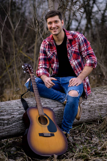 A handsome young man is sitting on a fallen tree next to an acoustic guitar in a nature park.