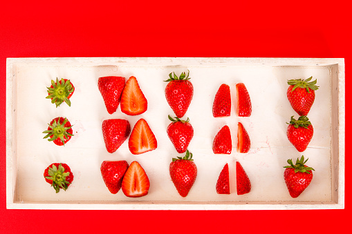 A number of strawberries, whole and sliced, on a wooden tray against a red background.