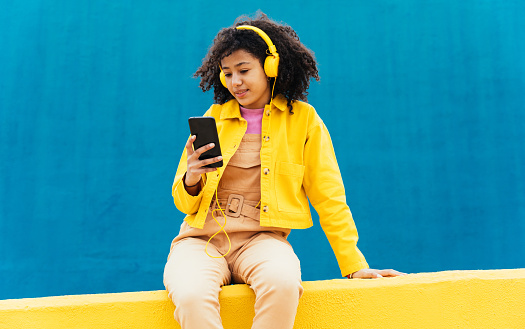 Young happy woman dancing and having fun outdoor. Teenager listening to music with smartphone and headphones in a yellow and blue modern urban area