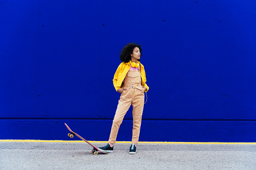 Young happy woman having fun outdoor with her skateboard. Teenager listening to music with smartphone and headphones in a yellow and blue modern urban area