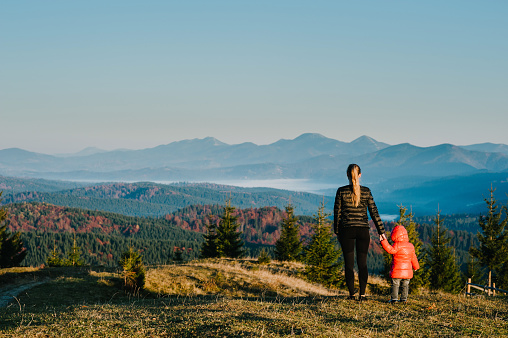 Mother and daughter, enjoying time together, stand on sunset on top of foggy mountain. Tourists on background autumn nature. Hikers on sunlight in country Europe. Happy family holidays. Back view.