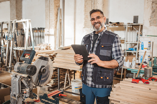 Carpenter in wood workshop using digital tablet for online buying or banking. Mature man in working uniform. Space is full of working tools and wooden planks.