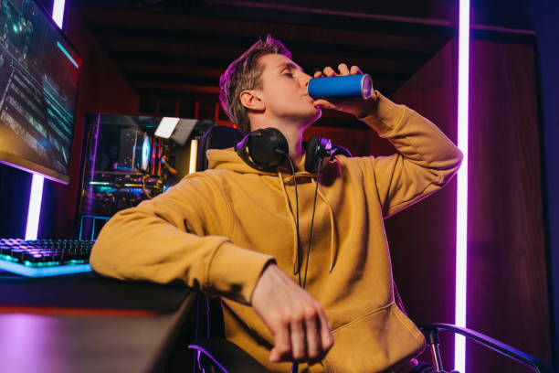 Yong caucasian pro gamer drinking energy drink while playing computer video games Portrait of young professional cyber sportsman sitting at gaming desk at home and drinking caffeine energy drink to concentrate on online video shooter game at night. Neon coloured room. Cyber sport energy drink stock pictures, royalty-free photos & images