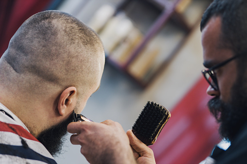 Hipster barber focused on grooming a beard in his barber shop. Retro barber shop. Attractive barber cutting hair. Man getting his beard groomed.