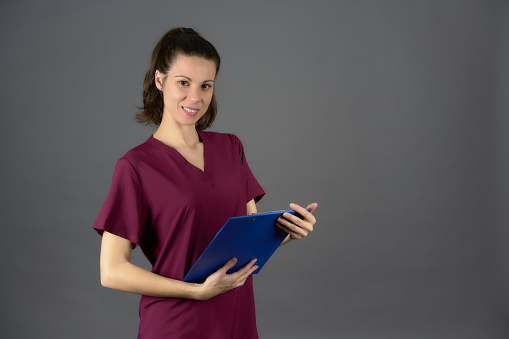 nurse in purple uniform looking into camera with medical report on a blue clipboard on a gray background with side copy space