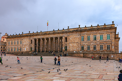 Bogota, Colombia - July 01, 2016: Looking across Plaza Bolivar, in the Andean capital city of Bogota in South America, to the building that is the official place of business for both houses of parliament: the Senate and the Congress. Completed in 1926, it was designed in the 19th Century by the Danish Architect Thomas Reed. Local Colombian people nonchalantly walk past the building of prime importance to the Country, as they go about their daily lives. There are a few international tourists in the image as well. The Colombian national flag flies over the building. There are pigeons all over the square looking for scraps of food. The square slopes from East on the left, to West on the right. The altitude at street level is 8,660 feet above mean sea level. Photo shot in the afternoon sunlight, on a cloudy, overcast day; horizontal format. Copy space.