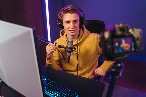 Young caucasian man pro gamer shooting video for his vlog on camera, talking on microphone. Cyber sportsman recording live stream for his followers while playing computer games on his PC at home