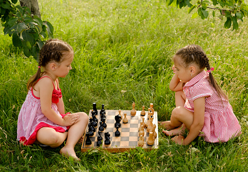 Children play chess sitting on the lawn. The kid has a thoughtful look. The girl thinks over her next move with a piece. Strategic Board Games