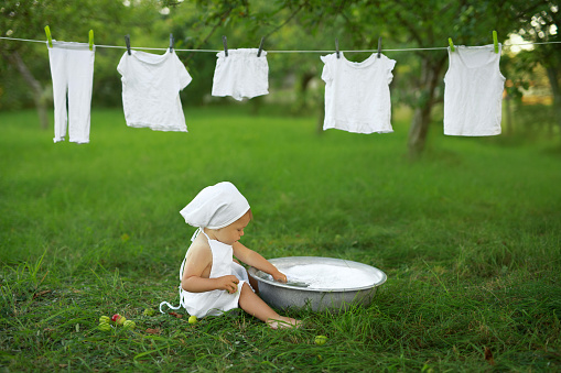 Baby washes white clothes in basin in the garden in nature. Girl in an apron and a kerchief is sitting on the lawn on grass. Baby Washing Hypoallergenic Powder Laundry Detergents Hand Washing Powders