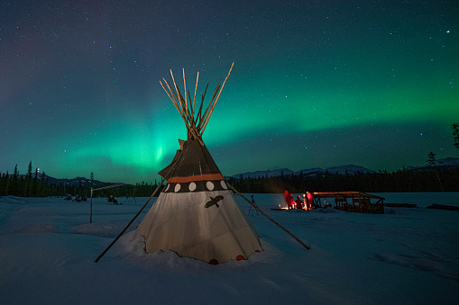 Group of people sitting around a campfire, and a teepee, under the northern lights in the Yukon
