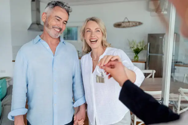 Real estate agent giving a mature couple the key to their new house. All are happy and smiling. The house can be seen in the background.